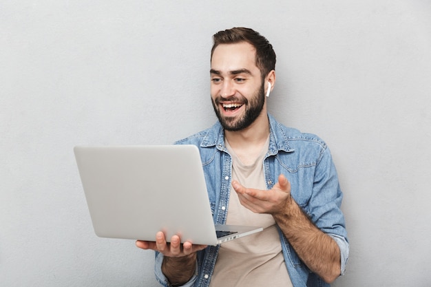 brunette lachende man met baard met laptop en met behulp van earpod geïsoleerd over grijze muur