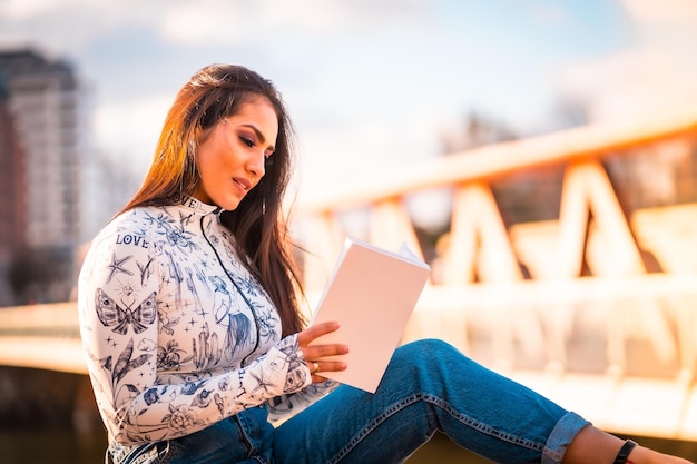 A brunette Latin girl in the city at sunset by the river enjoying reading a book
