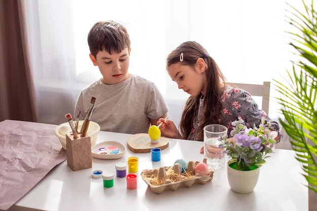 A brunette little girl and boy sits at a white table near the window paints eggs