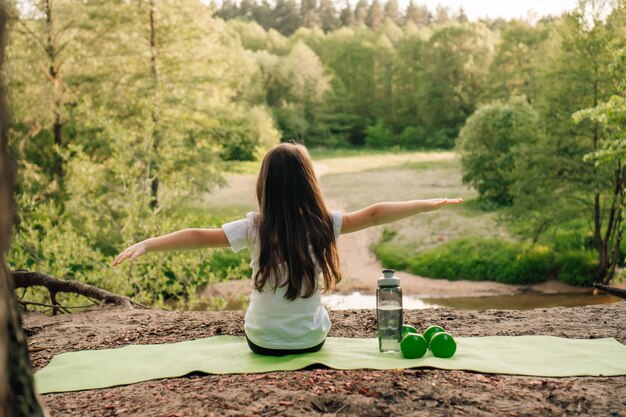 Foto ragazzina bruna con le braccia spalancate seduta su un tappeto verde su un terreno sabbioso manubri e bottiglia sdraiati vicino all'atleta