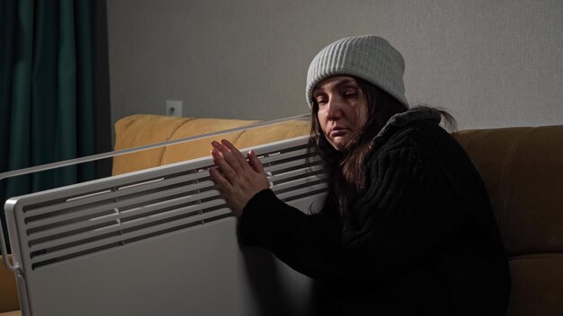 Brunette long-haired woman wearing knitted hat and sweater catches cold and sits on soft sofa embracing heater to feel warm in living room.