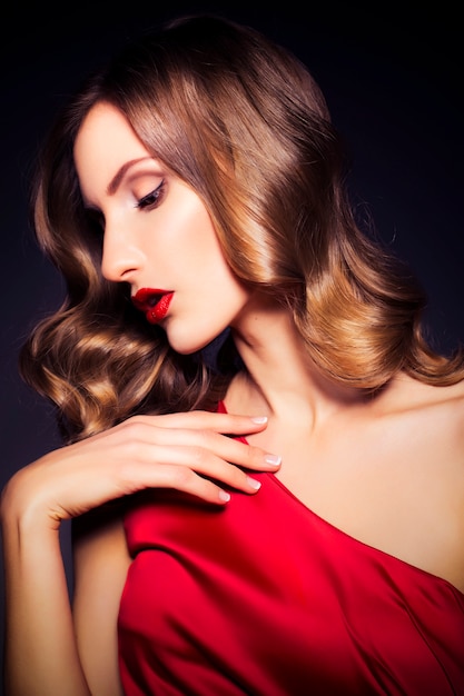 Brunette luxury woman in red dress with clear skin and evening dark make up: green cat eye and brown eyeshadows. Waved hairstyle. Dark background