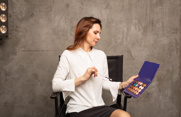 A brunette makeup artist in a white blouse is sitting on a chair with a palette of shadows on a gray background with a place for text. Beauty Specialist