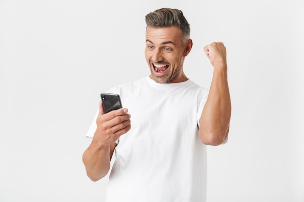 Brunette man 30s wearing casual t-shirt rejoicing while using smartphone isolated on white