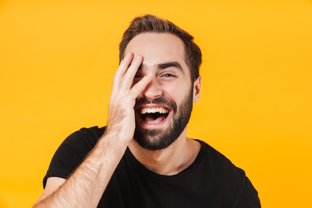 brunette man in basic black t-shirt laughing and touching his face isolated over yellow wall