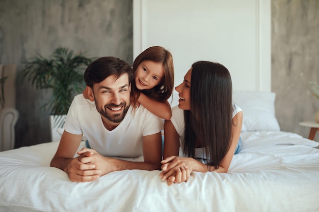 Brunette man laughing while his wife and daughter lying on a bed together with him