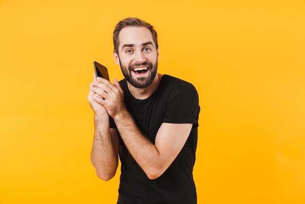 brunette man met een zwart basic t-shirt, blij en pratend op smartphone geïsoleerd over gele muur