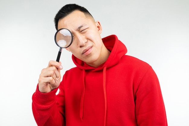 Photo brunette man in a red sweater looking at the camera through a magnifying glass on a white isolated background.