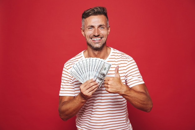 brunette man in striped t-shirt smiling and holding fan of money in cash isolated on red