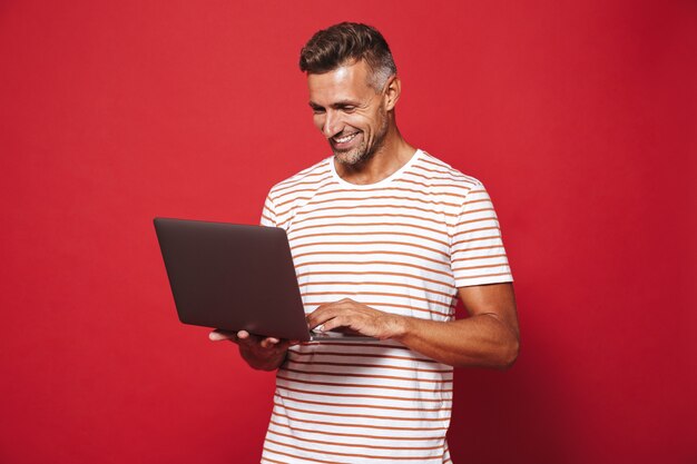 brunette man in striped t-shirt smiling and holding gray laptop isolated on red