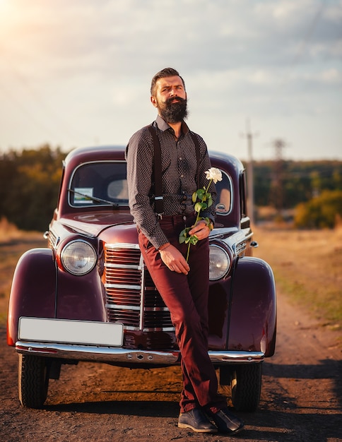 A brunette man with a thick black beard in a shirt, trousers with suspenders holds a white rose in his hand near a brown retro car on a rural road.
