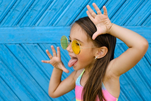 Foto brunette meisje in gele zonnebril staat tegen een blauwe muur en maakt een grappige grimas met tong