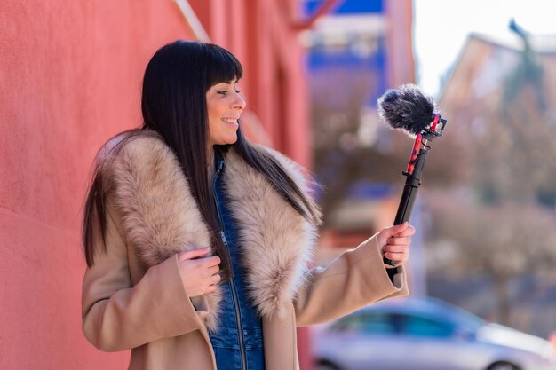 Brunette meisje lacht en zwaait tijdens het opnemen van een videoblog met haar mobiele telefoon en microfoon, gekleed in een winterjas voor sociale netwerken. Kaukasisch mooi een rode achtergrond