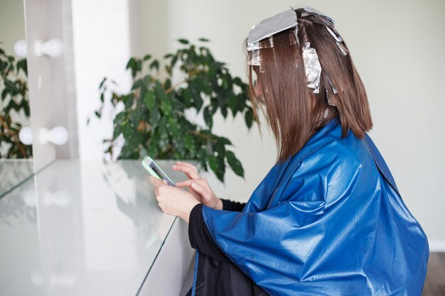 Brunette meisje verft haarlokken in de salon en kijkt naar zijn telefoon tijdens het wachten