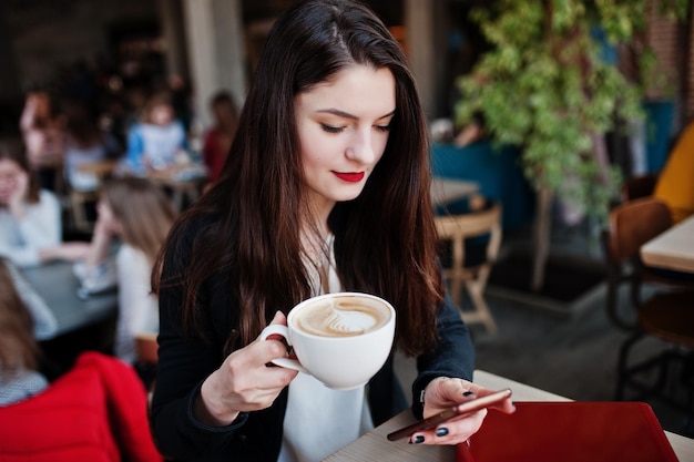 Brunette meisje zittend op café met kopje cappuccino werken met rode laptop en kijken naar mobiele telefoon