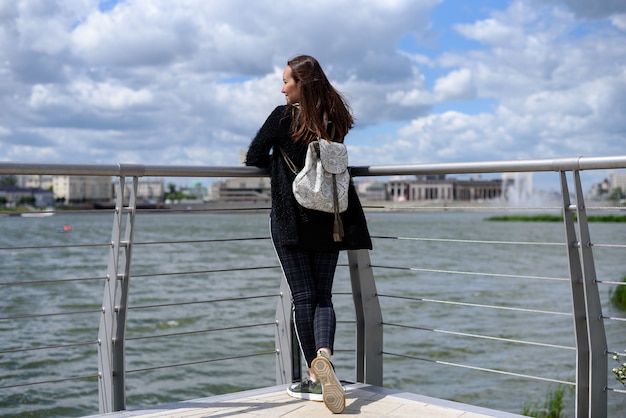 Brunette met een rugzak staat op het observatiedek bij het meer en bewondert het uitzicht