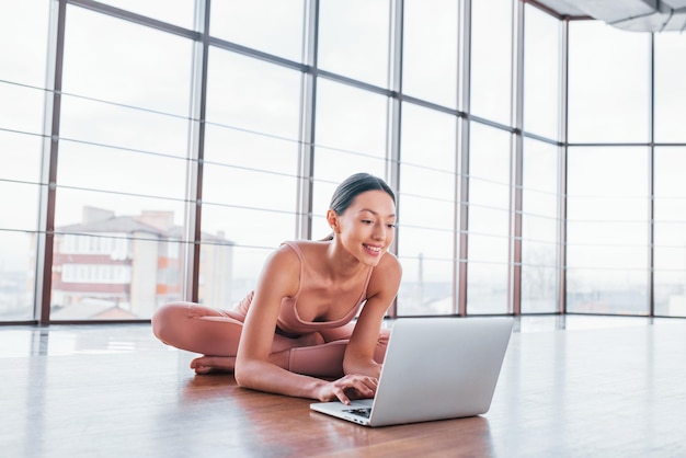 Brunette met slank lichaamstype die oefeningen op de vloer doet en laptop gebruikt.