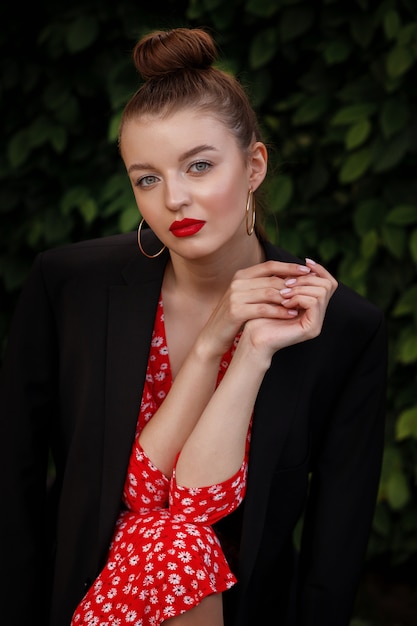 brunette model met verzameld haar en make-up in een rode jurk en zwarte heren jas zit in het park in de zomer.