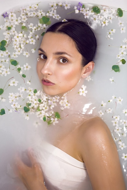 Brunette model woman lies in a white bath with milk and small flowers