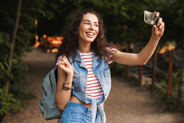 Brunette mooie vrouw met rugzak, lachen en selfie foto nemen op smartphone tijdens het wandelen langs pad in groen park