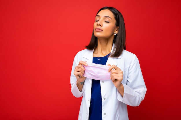 Brunette mooie vrouw opstijgen virus beschermend masker op gezicht tegen coronavirus en witte medische jas geïsoleerd op rode achtergrond.