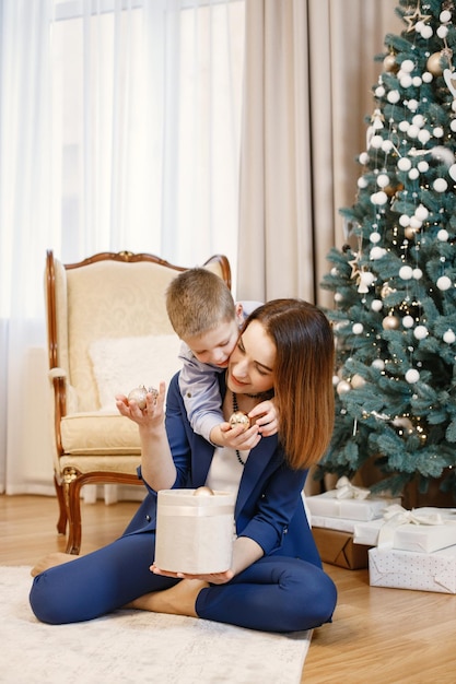 Brunette mother and little son sitting on the floor near christmas tree at home. Family is going to decorate Christmas tree together with balls. Woman wearing blue costume and boy blue shirt.