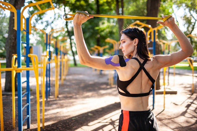 Brunette muscular woman posing with fitness resistance band in park, sports ground on