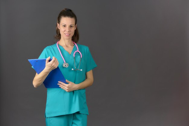 Brunette nurse smiling straight ahead holding a clipboard with a pink stethoscope a gray background