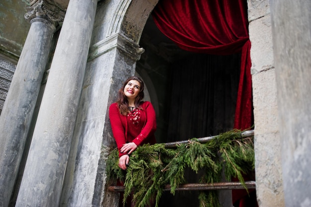 Brunette plus size model at red against arch with curtains.