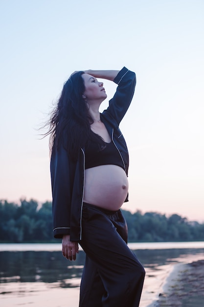 Brunette poses against background of nature attractive young pregnant woman in dark silk pajamas pos...