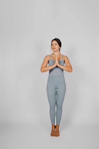 Brunette poses on isolated background Young woman in gray yoga tracksuit performs stretching exercise she stands on her toes and put her palms together concept of healthy lifestyle