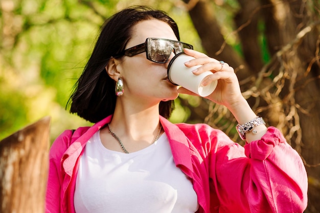 コーヒーカップを保持し、公園を歩いているサングラスのブルネットのきれいな女性