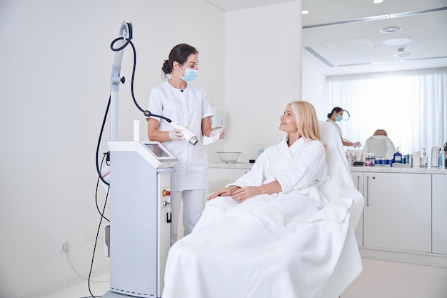 Brunette professional doctor in medicine uniform and mask talking with patient in beauty clinic