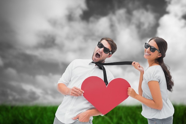 Photo brunette pulling her boyfriend by the tie holding heart against green grass under grey sky