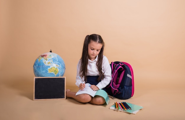 A brunette schoolgirl in a uniform is sitting with school supplies and writing in a notebook on a beige background with a place for text