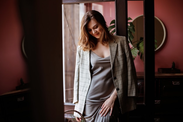 Brunette in a silk dress and jacket stands by the window in the apartment