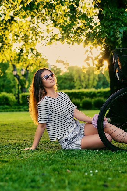 Brunette sits on the green lawn with bicycle and holds flower bouquet in park.