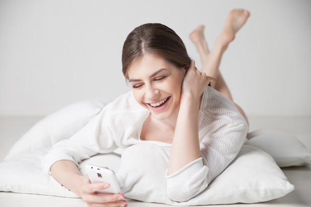 Brunette smiling while using smartphone on pillow at home