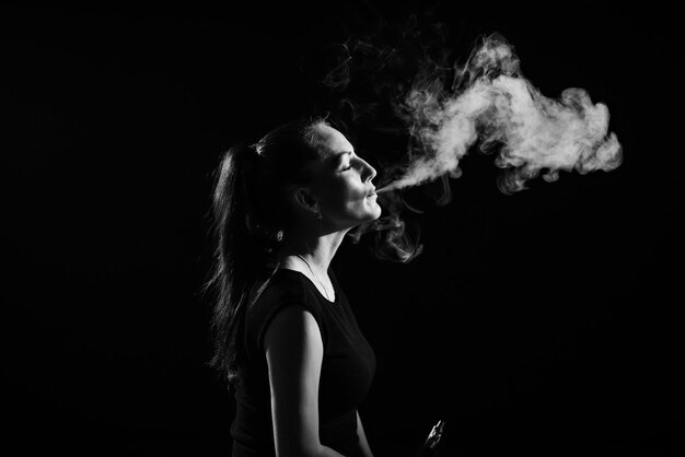 The brunette smokes an electronic cigarette on a black background releases thick smoke from her mouth Monochrome Portrait of a woman smoking a vape hovering