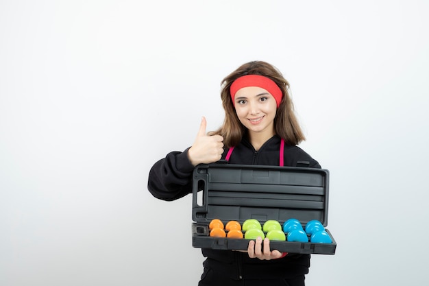 Brunette sportswoman holding colorful dumbbells and showing thumb up .