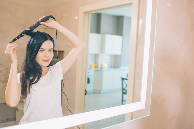Brunette stands at mirror and using curler hair. She is in bathroom. Woman holds curler with both hands. She makes hair dressing.