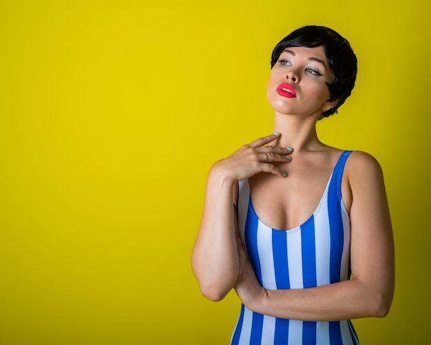 Brunette in a striped swimsuit posing in the studio on a bright yellow background Woman in a short wig with sensual lips