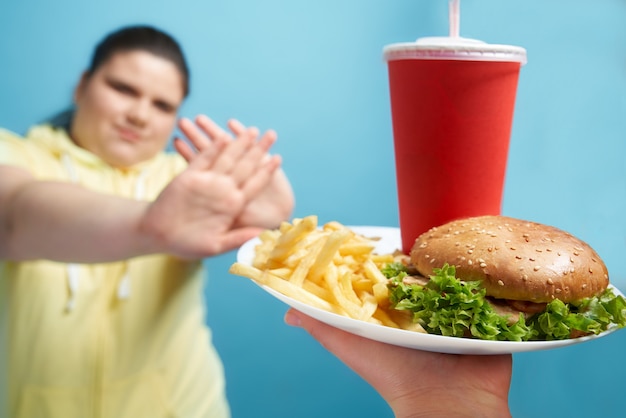 Brunette te dikke vrouw met gesloten ogen, armen gebouwd als een kruis om te laten zien dat ze geen fastfood op een witte plaat wil eten. Jonge mooie dame maakt de juiste keuze voor haar gezonde levensstijl