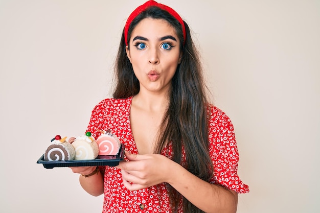 Brunette teenager girl holding cake sweets scared and amazed with open mouth for surprise disbelief face