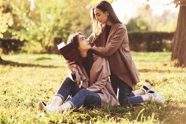 Brunette twin girls sitting on the grass and looking at each other. One of them is holding brown book, onother one is leaning on sisters shoulders from the back in autumn park on blurry background.