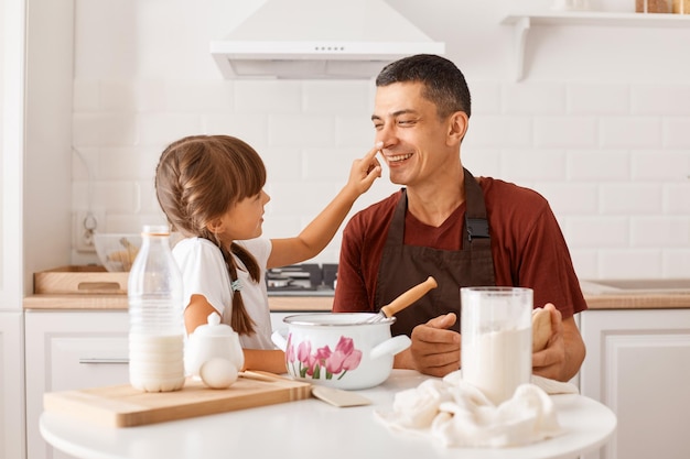 Brunette vader draagt een bordeauxrood t-shirt en bakt samen met zijn vrouwelijk kind in de keuken terwijl hij aan tafel zit, dochter smeert de neus van de vader in met bloem, familie heeft plezier tijdens het koken.
