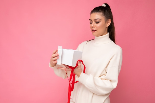 brunette vrouw geïsoleerd op roze achtergrond muur dragen witte trui bedrijf geschenkdoos en unboxing aanwezig camera kijken.