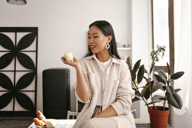 Brunette vrouw in beige vestbroek zit op bureau en kijkt naar appel Gelooide Aziatische dame in stijlvolle outfit glimlacht op keuken