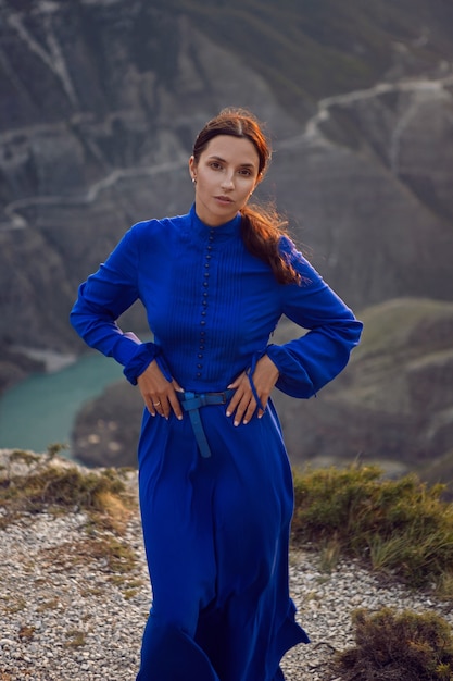 Brunette vrouw in een blauwe lange jurk staat 's avonds bij zonsondergang aan de rand van de Sulak-canyon in Dagestan