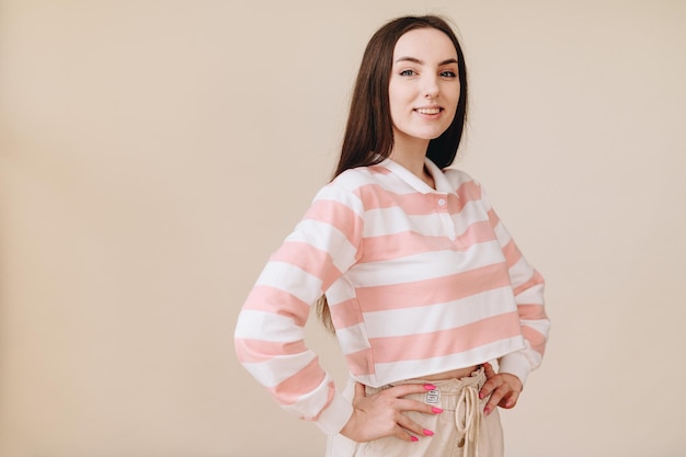 Brunette vrouw in vrijetijdskleding op beige achtergrond staat zijdelings handen op haar heupen glimlacht breed